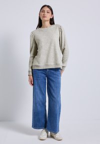 Pullover grigio chiaro con maniche a sbuffo, tessuto testurizzato e piccoli dettagli decorativi. Abbinato a jeans in denim blu a gamba larga e scarpe da ginnastica.