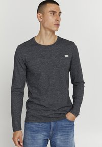Dunkelgraues Langarmshirt mit rundem Halsausschnitt, strukturiertem Stoff und einem kleinen weißen Markenlabel auf der Brust. Kombiniert mit blauen Jeans.