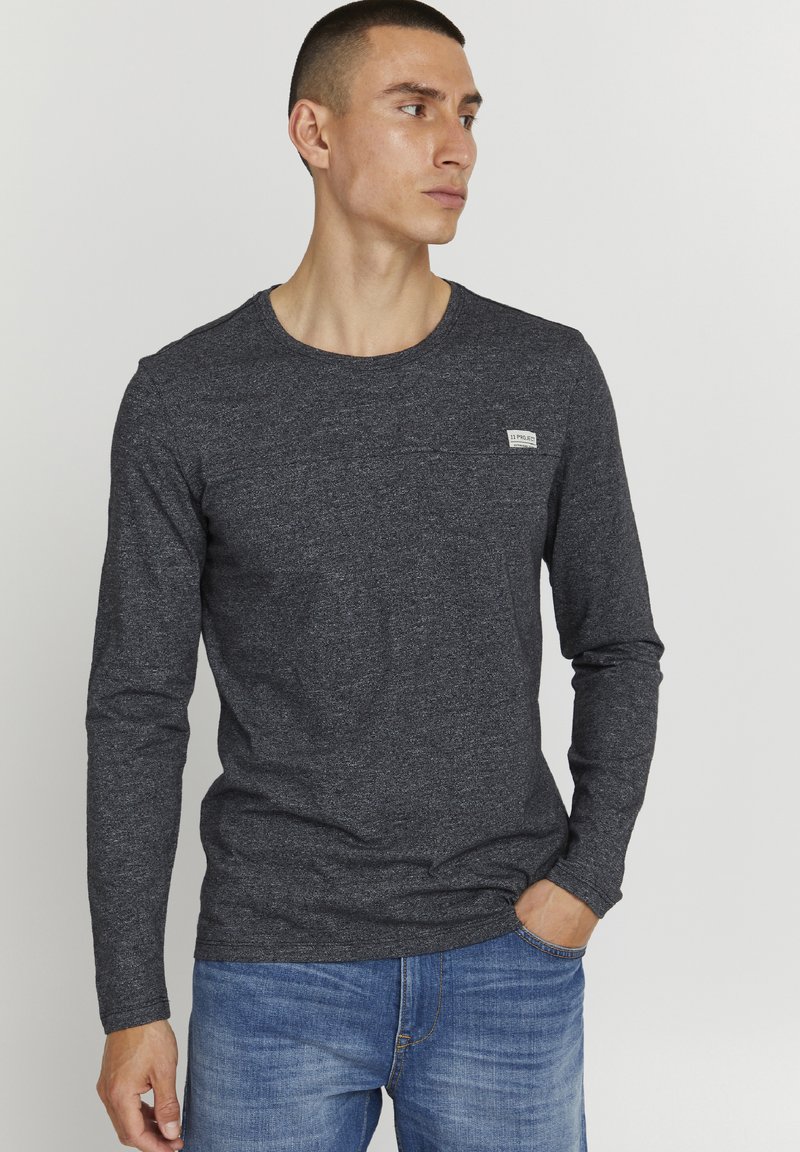 Dunkelgraues Langarmshirt mit rundem Halsausschnitt, strukturiertem Stoff und einem kleinen weißen Markenlabel auf der Brust. Kombiniert mit blauen Jeans.
