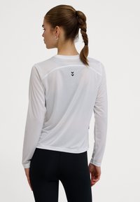Langärmliges, weißes Sportoberteil mit leichtem Mesh-Material, versehen mit schwarzen Chevron-Akzenten auf dem Rücken und einem lockeren Schnitt.