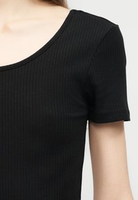 Κοντινό πλάνο ενός ατόμου που φορά μαύρο ριμπ πλεκτό τοπ με κοντά μανίκια και στρογγυλή λαιμόκοψη, μπροστά σε απλό φόντο.