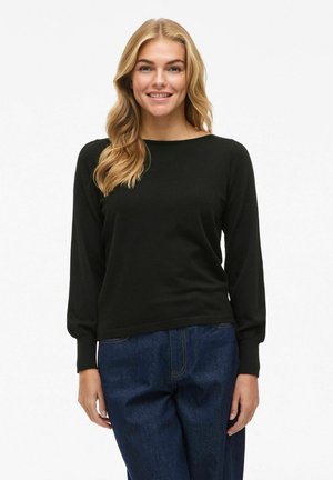 Donna sorridente con lunghi capelli biondi, che indossa un top nero a maniche lunghe e jeans blu, in piedi contro uno sfondo bianco uniforme.