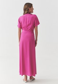 Fuchsia maxi dress with short sleeves and a fitted waist, featuring a smooth texture and flowing design. A patterned scarf is tied at the neck.