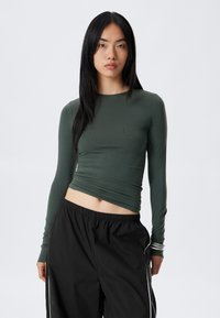 Long-sleeve, fitted top in dark green, made of smooth fabric. Features a crew neckline and a cropped cut. Paired with black wide-leg trousers.
