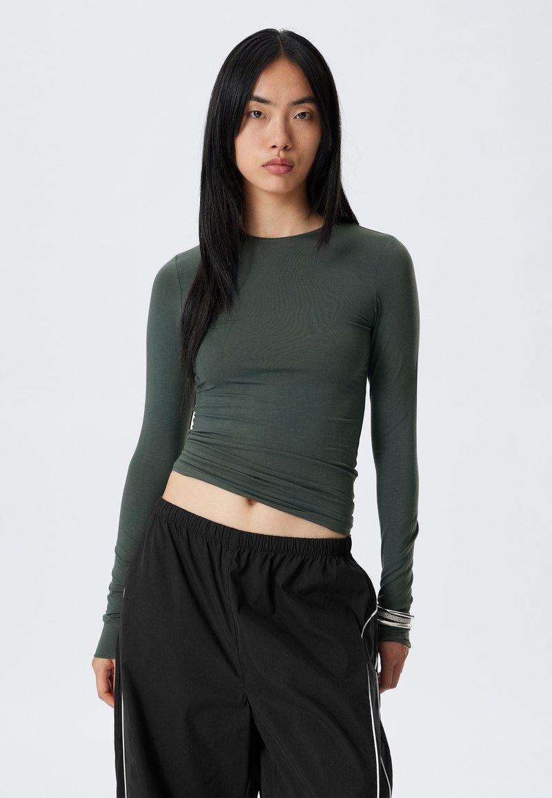 Long-sleeve, fitted top in dark green, made of smooth fabric. Features a crew neckline and a cropped cut. Paired with black wide-leg trousers.
