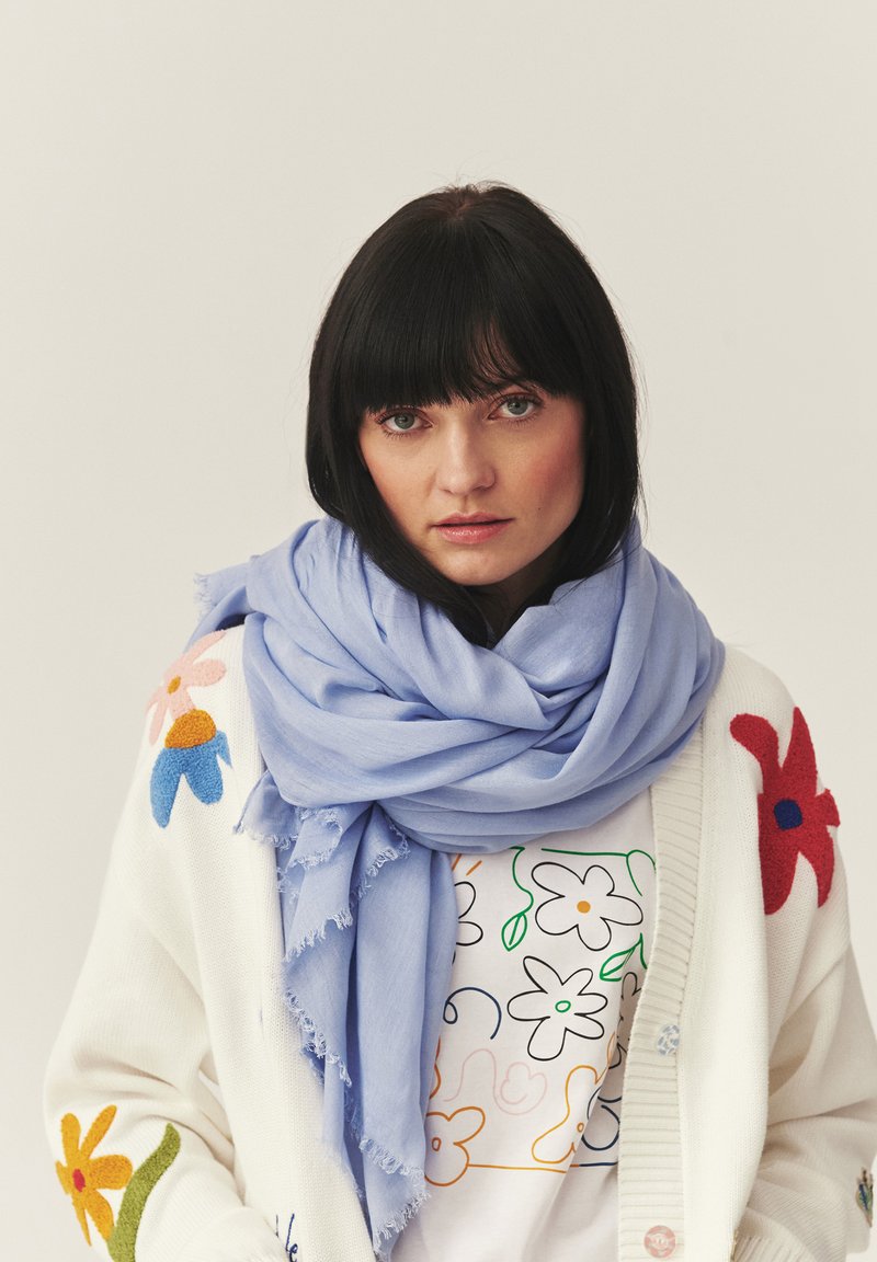 Femme aux cheveux foncés portant une écharpe bleu clair, un cardigan blanc avec une broderie florale colorée et une chemise à motif floral, regardant devant elle.