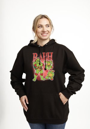 Femme souriante portant un sweat à capuche noir avec trois illustrations des Tortues Ninja adolescents et le texte « RAPH » sur le devant.