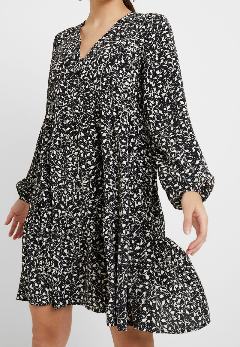 Robe à manches longues noir et blanc avec un motif floral et à pois, encolure en V, et une jupe ample et fluide portée par une femme.