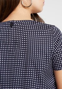 Blouse bleu marine avec un motif en grille composé de petits carrés blancs, col rond et fermeture à boutons dans le dos. Tissu à texture lisse.