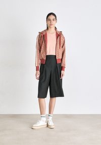 Hettejakke i burgunder med mønster, rosa t-skjorte, svarte plisserteshorts, hvite joggesko med beige såler og lyse stripete sokker.