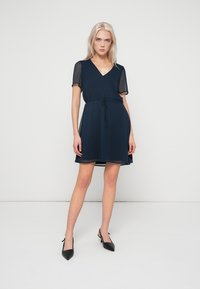 Σκούρο μπλε φόρεμα με κοντά διαφανή μανίκια, V-λαιμόκοψη και ζώνη στη μέση. Διαθέτει φουσκωτή φούστα που φτάνει πάνω από τα γόνατα. Μινιμαλιστικός σχεδιασμός.