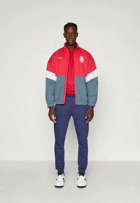 Mann steht da und trägt eine rot-weiße-graue Jacke mit Logo, ein rotes T-Shirt, marineblaue Hosen und weiße Sneakers vor einem einfarbigen Hintergrund.