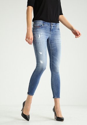 Femme portant un jean skinny bleu clair déchiré et des talons hauts noirs, debout devant un fond clair uni.