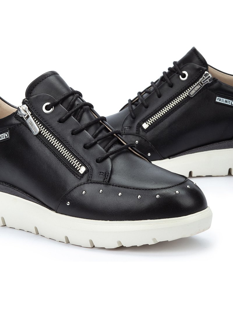 Zapatos de cuero negro con detalle de cremallera de metal, parte delantera con cordones y adornos de tachuelas plateadas; cuentan con una suela blanca gruesa para mayor comodidad.