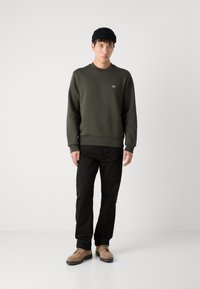 Felpa verde oliva con scollo a girocollo, polsini e orlo a coste. I pantaloni neri e le scarpe in camoscio color sabbia completano l'outfit. Indossato un berretto nero.
