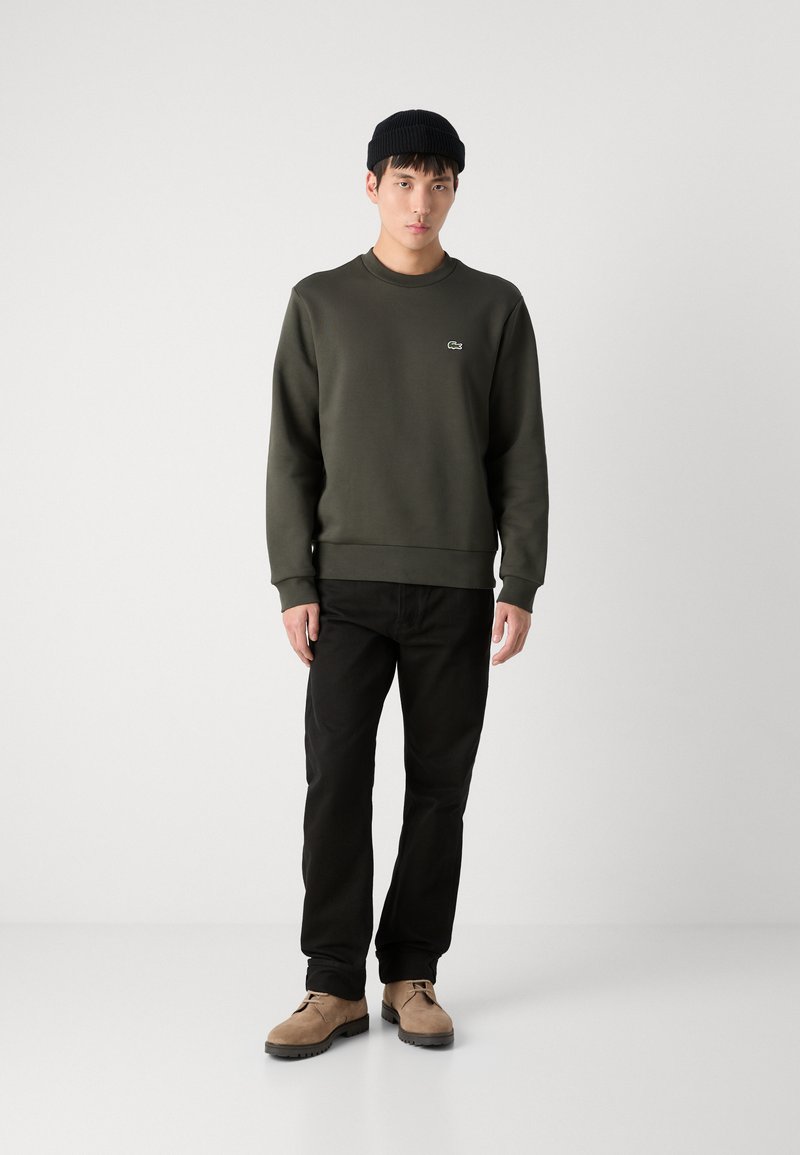 Sweatshirt vert olive avec col rond, poignets et ourlet côtelés. Un pantalon noir et des chaussures en daim tan complètent la tenue. Un bonnet noir est porté.
