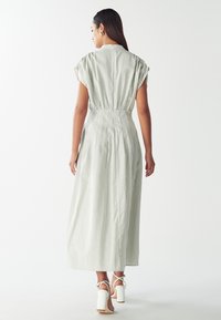 Maxi šaty v světle zelené barvě s bílými vertikálními pruhy; nabíraný pas, krátké rukávy a splývavá látka; kombinované s bílými podpatky.