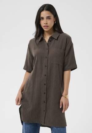 Kobieta o ciemnych włosach, ubrana w luźną, brązową sukienkę koszulową zapinaną na guziki oraz niebieskie jeansy na jednolitym jasnym tle.