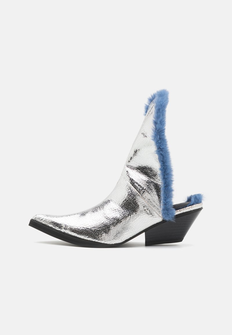 Silberne metallische Knöchelschuh mit spitzem Absatz, ausgestattet mit einem blauen Fellkragen und einem niedrigen schwarzen Keilabsatz. Die strukturierte Oberfläche reflektiert das Licht.