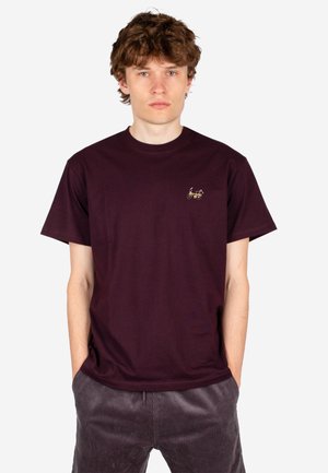 Junger Mann mit lockigem Haar, der ein dunkelviolettes T-Shirt mit einer kleinen Fahrrad-Stickerei auf der linken Brust und dunkle Hosen trägt.