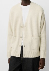 Beige, stickad kofta med v-ringning, fem knappar och två framfickor. Ribbad mudd och nederkant ger textur. Bärs över en vit t-shirt.