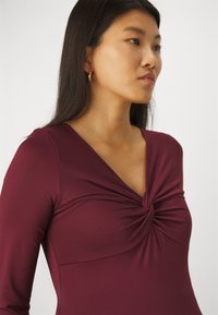 Burgundy long-sleeve top with a twisted knot detail at the neckline, made from smooth, stretchy fabric, featuring a v-shaped collar.