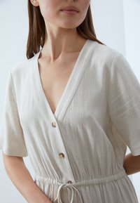 Lys beige lin kjole med V-hals, korte ermer og snøring i livet. Har knappdetaljer og strukturert stoff.