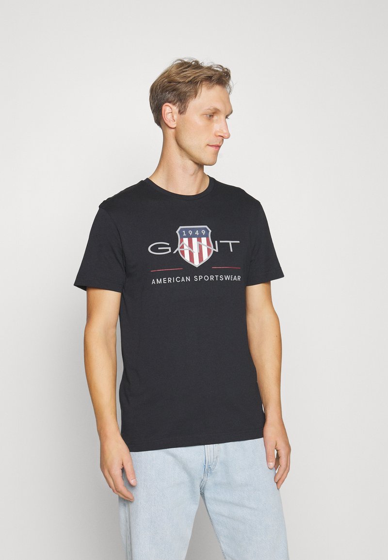 Svart bomull T-skjorte med logo foran med et skjolddesign i rødt og blått, korte ermer, rund halslinning og løs passform.