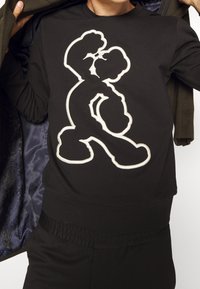 Felpa nera con una grafica contornata bianca di un personaggio dei cartoni animati. Collo rotondo, polsini e vita a costine con una texture liscia.