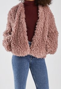 Luddig, kort kappa i ljusrosa, med en mjuk textur, överdimensionerade ärmar och en öppen frontdesign. Kombinerad med en ribbad vinröd topp och denimjeans.