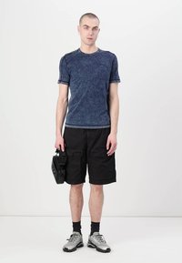 Tricou din denim albastru cu mânecă scurtă, pantaloni scurți negri tip cargo, adidași gri, ținând o geantă mică neagră. Design simplu, athletic, cu caracteristici practice.