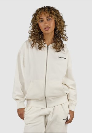 Vrouw met krullend haar, gekleed in een crèmekleurige sweater met ritssluiting en bijpassende broek, met zwarte "PEGADOR"-logo's op de borst en dij.