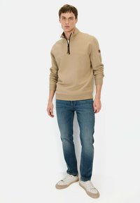 Beige kwart-rits trui met gestructureerde stof, geribbelde manchetten en een klein logo-accent. Gecombineerd met blauwe spijkerbroeken en witte sneakers.