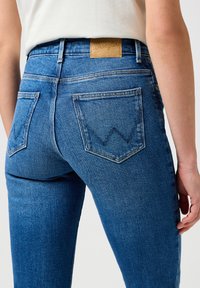Jean en denim bleu à taille haute, avec une étiquette en cuir portant le texte "Wrangler" et deux poches arrière avec un motif de couture distinctif.