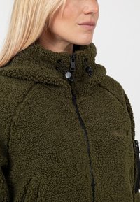 Oliwkowa polarowa kurtka z wysokim kołnierzem, z zamkiem błyskawicznym i regulowanymi trokami. Teksturowana powierzchnia oraz minimalny detal z logo.