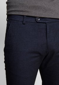 Pantalon bleu marine en tissu texturé, doté d'une fermeture à bouton et de poches latérales, avec une coupe slim.