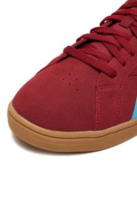 Nahaufnahme eines roten Wildledersneakers mit roten Schnürsenkeln, Gumsohle und blauem Detail an der Seite vor weißem Hintergrund.