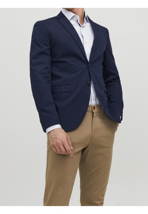Uomo con blazer blu navy, camicia bianca e pantaloni beige, in piedi con una mano in tasca su uno sfondo semplice.