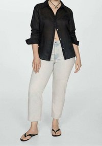 Camisa de lino negra con botones y mangas enrolladas, combinada con jeans de mezclilla de color claro y sandalias negras. Fondo neutro.