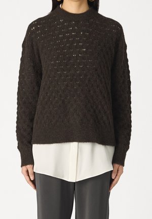 Femme portant un pull tricoté texturé marron foncé par-dessus une chemise blanche boutonnée, associé à un pantalon gris foncé coupé à la taille.