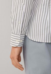 Weißes Hemd mit grauen vertikalen Streifen, Knöpfen an den Manschetten und abgerundetem Saum. Der strukturierte Stoff bietet einen subtilen Kontrast zur glatten Oberfläche.