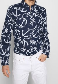 Chemise bleu marine à boutons avec motifs de croix et de corde blancs ; col classique et manches longues, assortie avec un jean blanc.