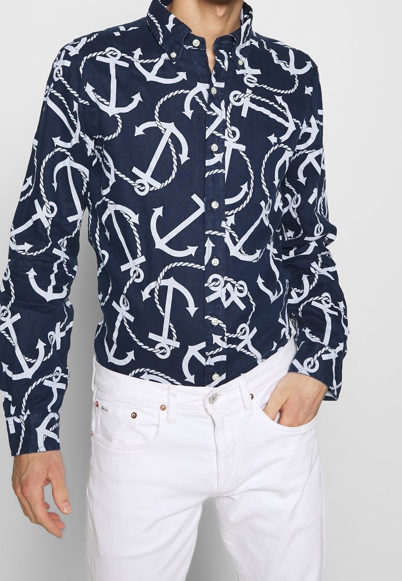 Chemise bleu marine à boutons avec motifs de croix et de corde blancs ; col classique et manches longues, assortie avec un jean blanc.
