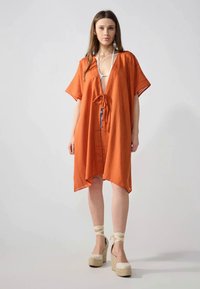 Cubierta de estilo kimono naranja con mangas cortas, lazo frontal y ajuste suelto; combinada con sandalias de plataforma de esparto beige.