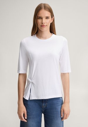 Vrouw met steil lichtbruin haar draagt een eenvoudig wit T-shirt met korte mouwen en een blauwe spijkerbroek, staand tegen een neutrale achtergrond.