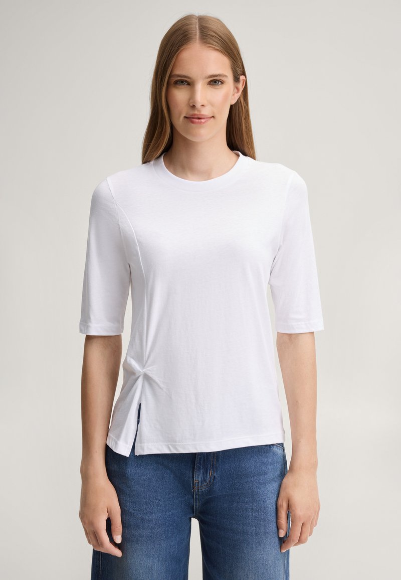 Femme aux cheveux lisses châtain clair porte un simple t-shirt blanc à manches courtes et un jean bleu, debout sur fond neutre.