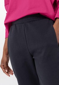 Tmavě šedé tepláky s měkkou texturou a elastickým pasem, spárované se světle růžovým topem s dlouhými rukávy. Na nohách je viditelná ruka, která spočívá na teplákách.