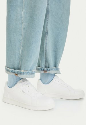 Hellblaue Jeans, an den Knöcheln hochgekrempelt, kombiniert mit hellblauen Socken und weißen Kappa-Sneakern auf weißem Hintergrund.