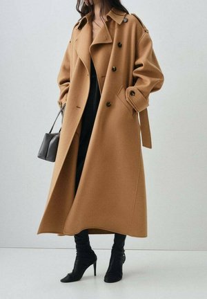 Femme portant un long manteau camel croisé, pantalon noir, bottes noires pointues à talons hauts, tenant un petit sac à main gris.