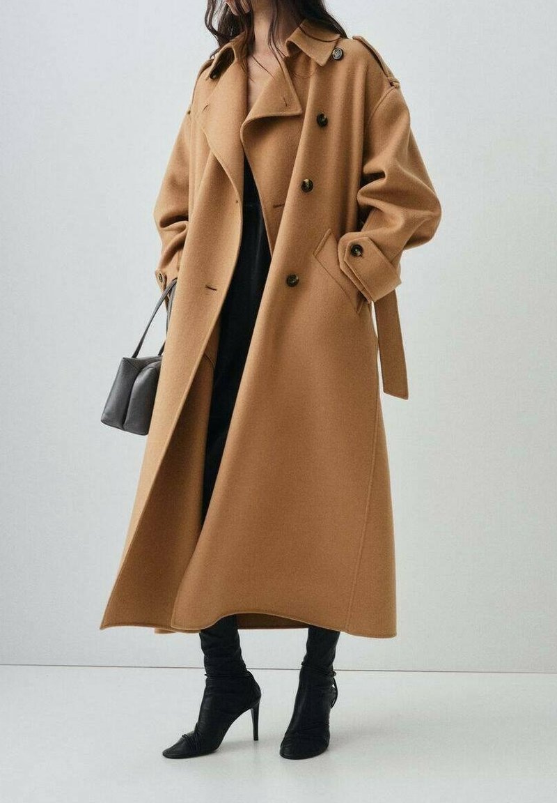 Femme portant un long manteau camel croisé, pantalon noir, bottes noires pointues à talons hauts, tenant un petit sac à main gris.